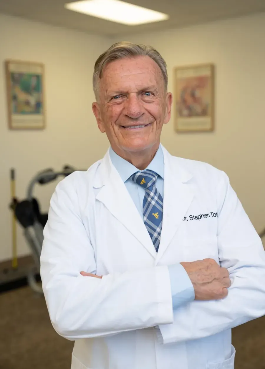 Dr. Stephen G. Toth smiling in his white coat, arms folded, in his Morgantown office